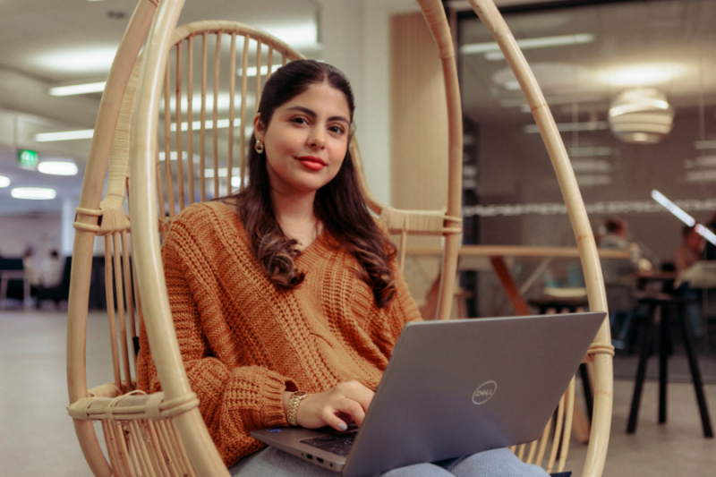 A student with a laptop sitting in a wicker chair at the university, smiling at the camera